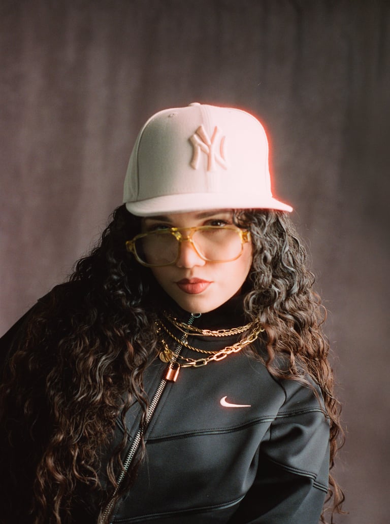 Person wearing a white Yankees cap, yellow-tinted glasses, and black Nike jacket with gold chains against a gray background