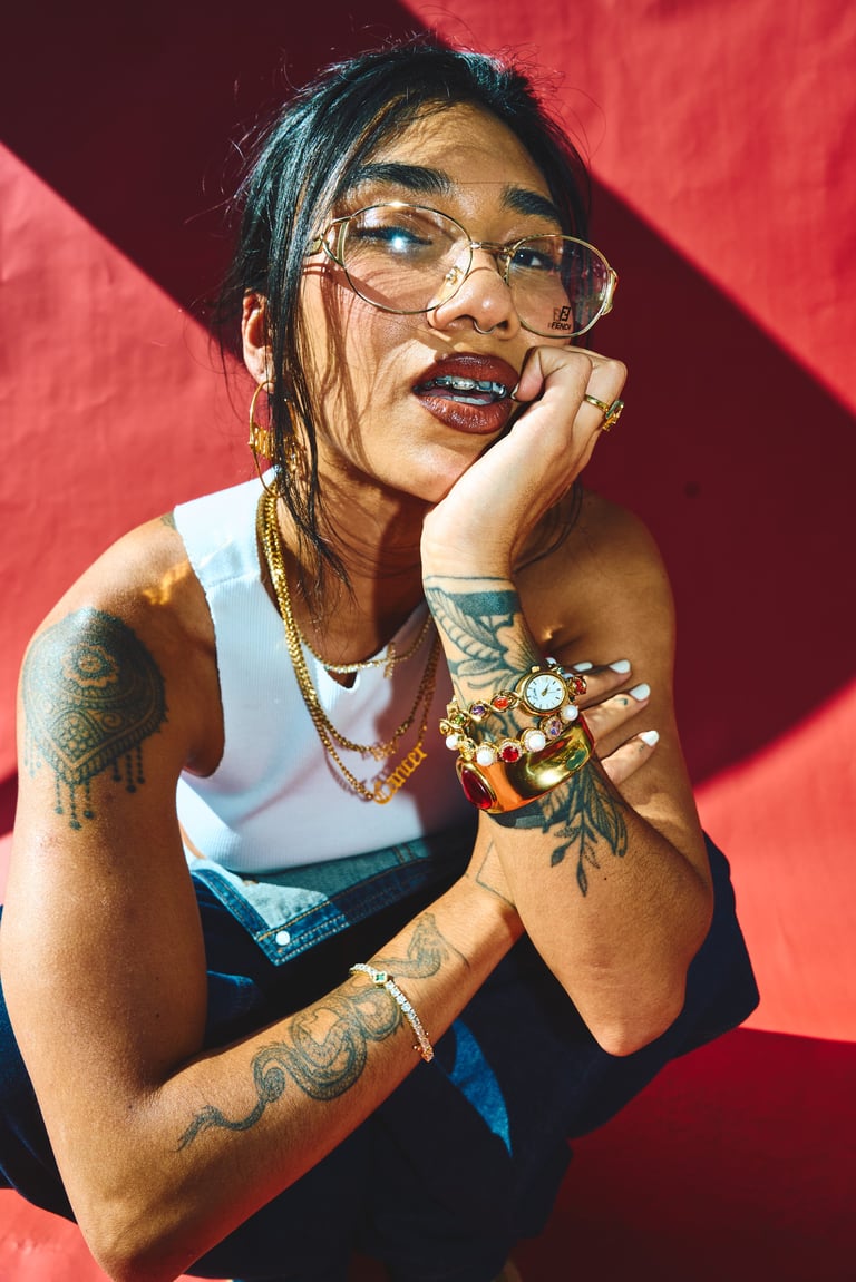 Person with dark braided hair and glasses wearing white tank top and jewelry, posing thoughtfully with hand on face against red background